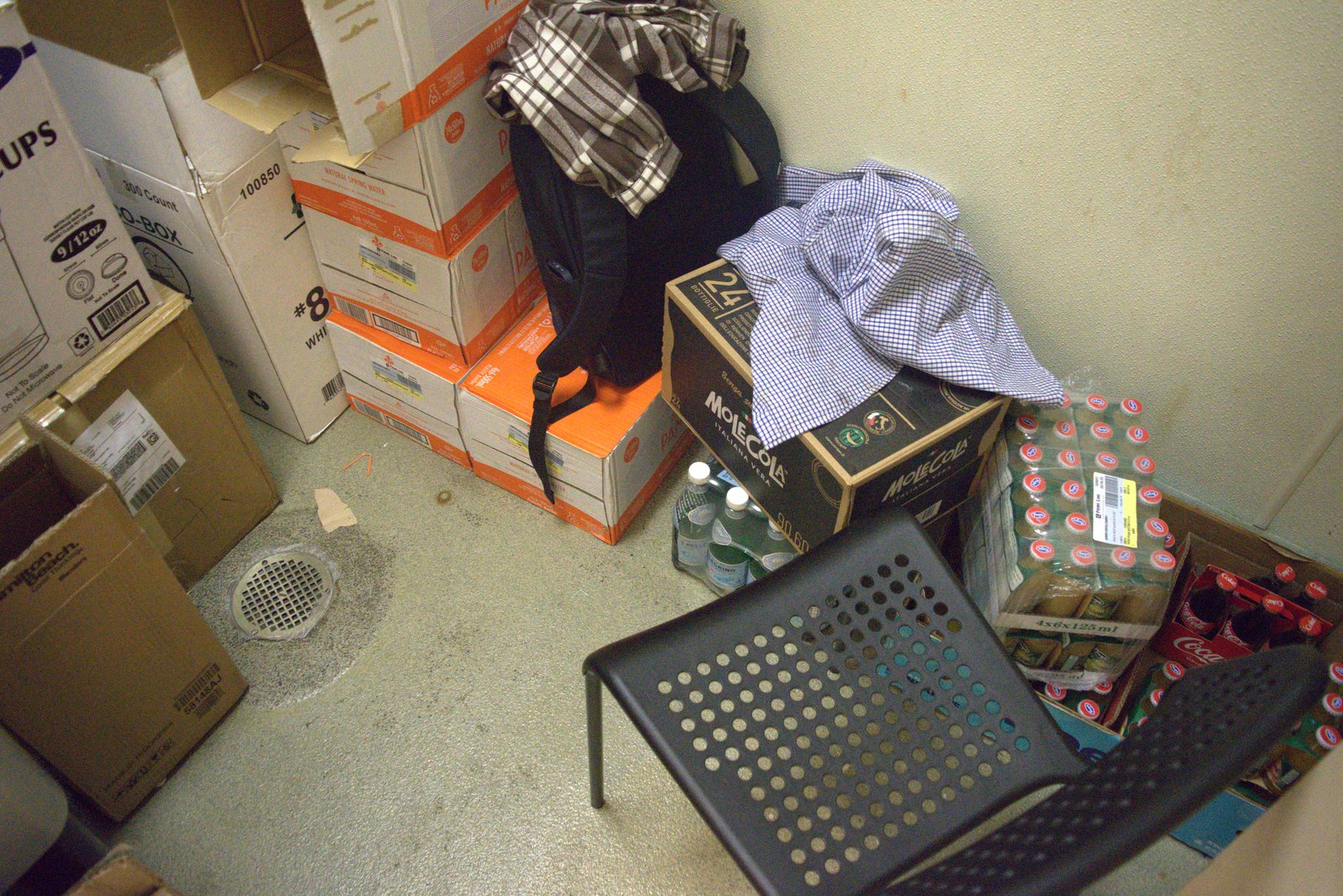 Restroom floor — Mole Cola cases, San Pellegrino, Coca-Cola and personal clothing piled next to a floor drain