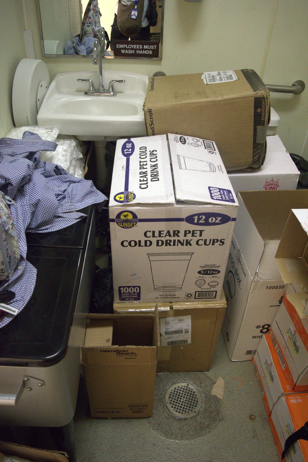 Hand-wash sink in employee restroom completely blocked by stacked cup boxes and personal laundry — basic hygiene impossible