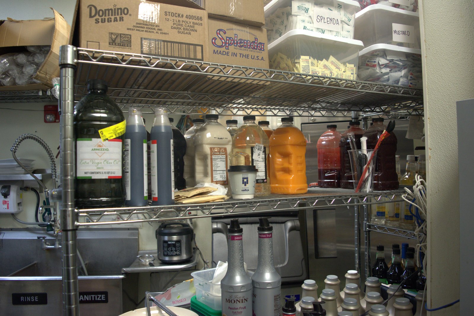 Back-of-house syrup shelving — unapproved Arrezzio olive oil, generic syrups and bulk Domino sugar mixed above a Lavazza-branded cup