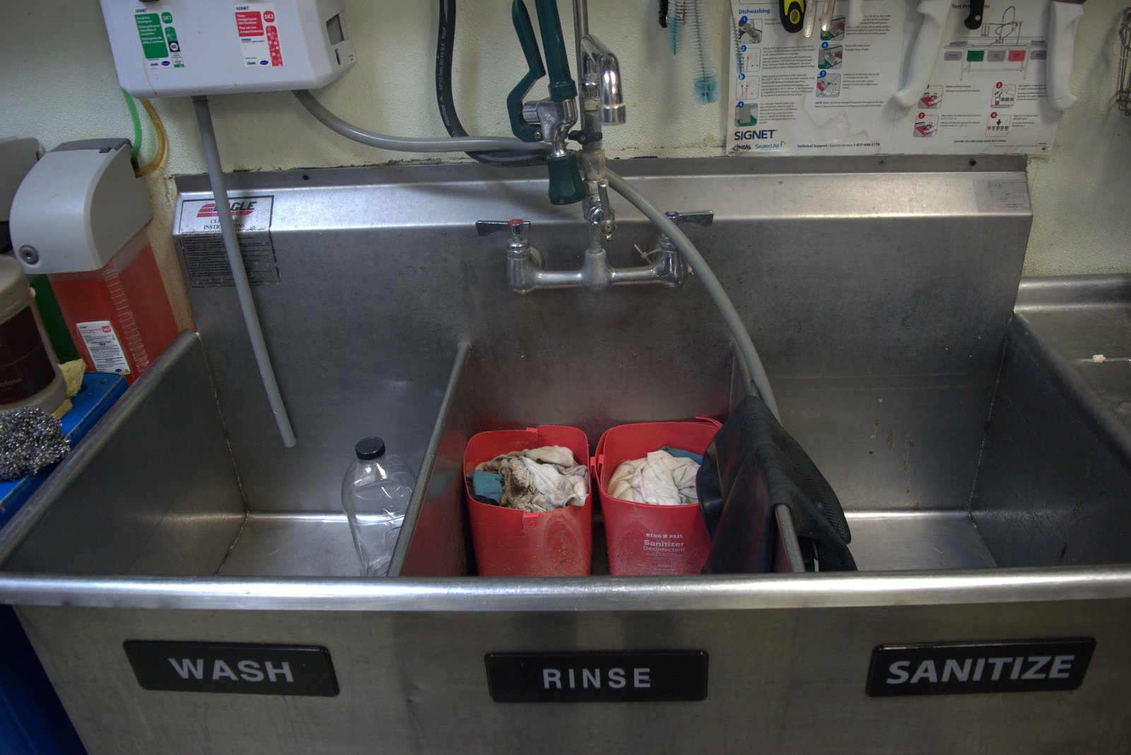 Three-compartment sink (Wash/Rinse/Sanitize) misused — sanitizer buckets full of soiled rags and a plastic bottle dumped in the wash basin