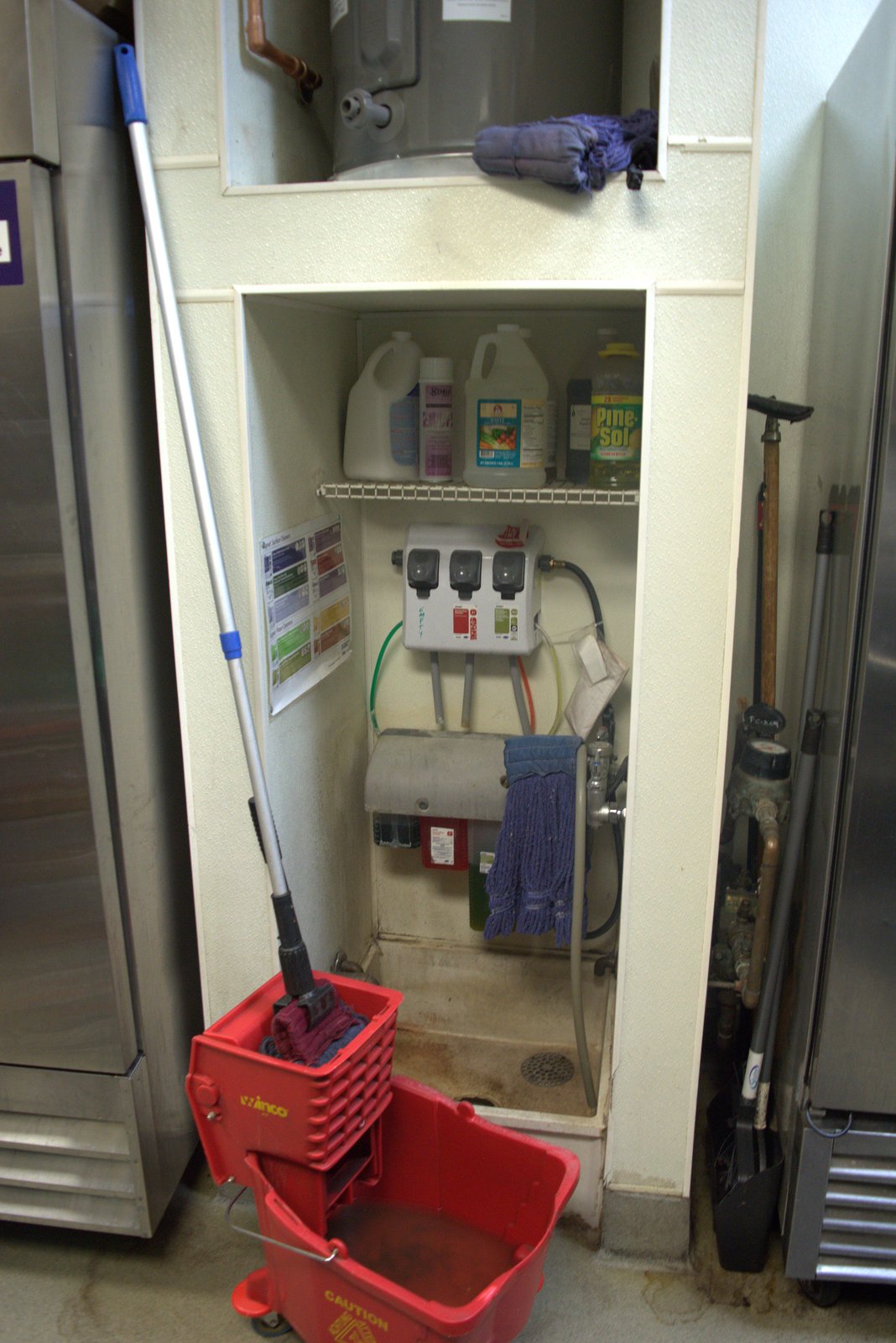 Mop closet — chemical bottles stored on a single open shelf above the mop sink with a soiled mop; no chemical segregation