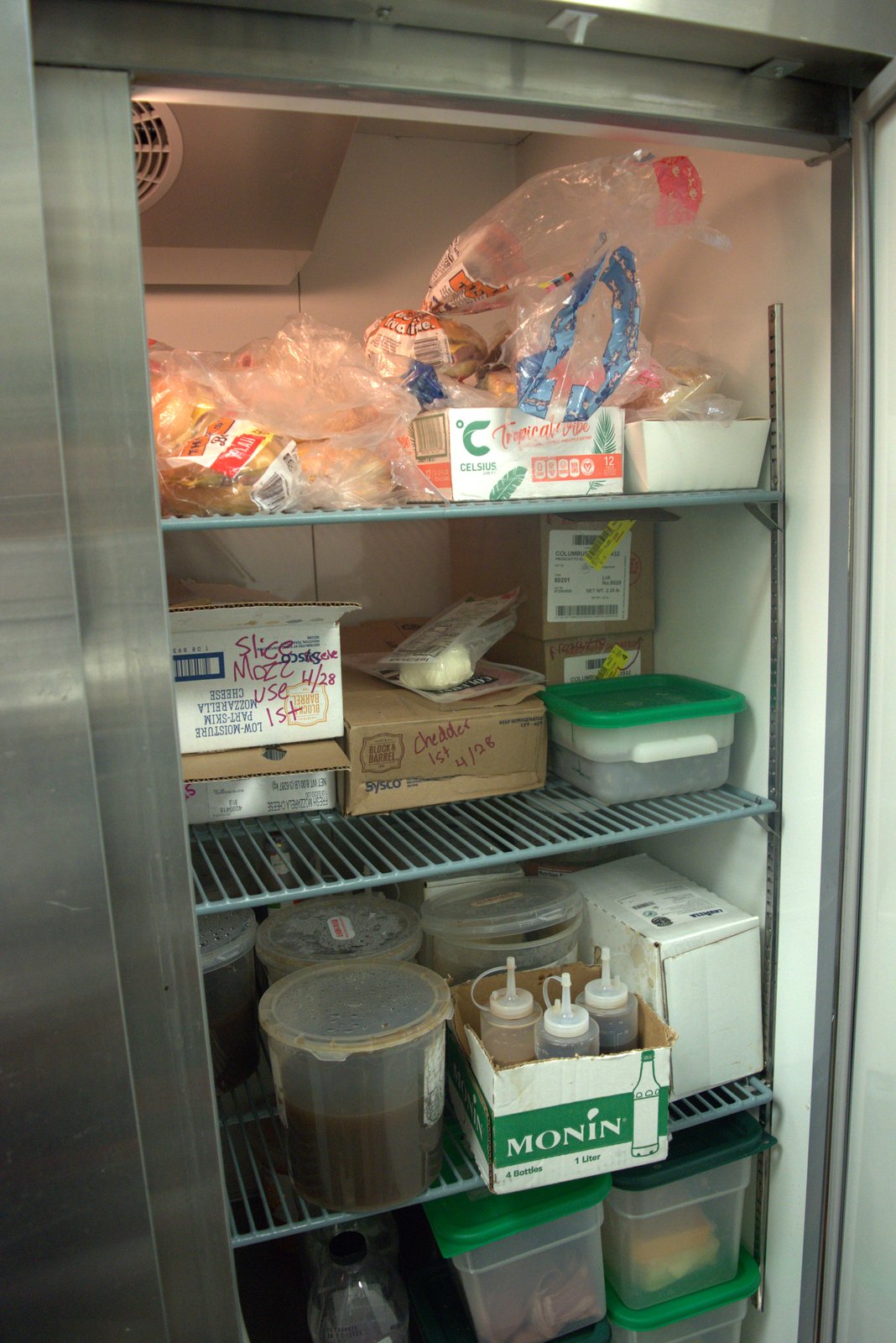 Reach-in fridge — handwritten 'Use 1st 4/28' on cheese boxes next to a Celsius energy-drink case (unauthorized SKU)