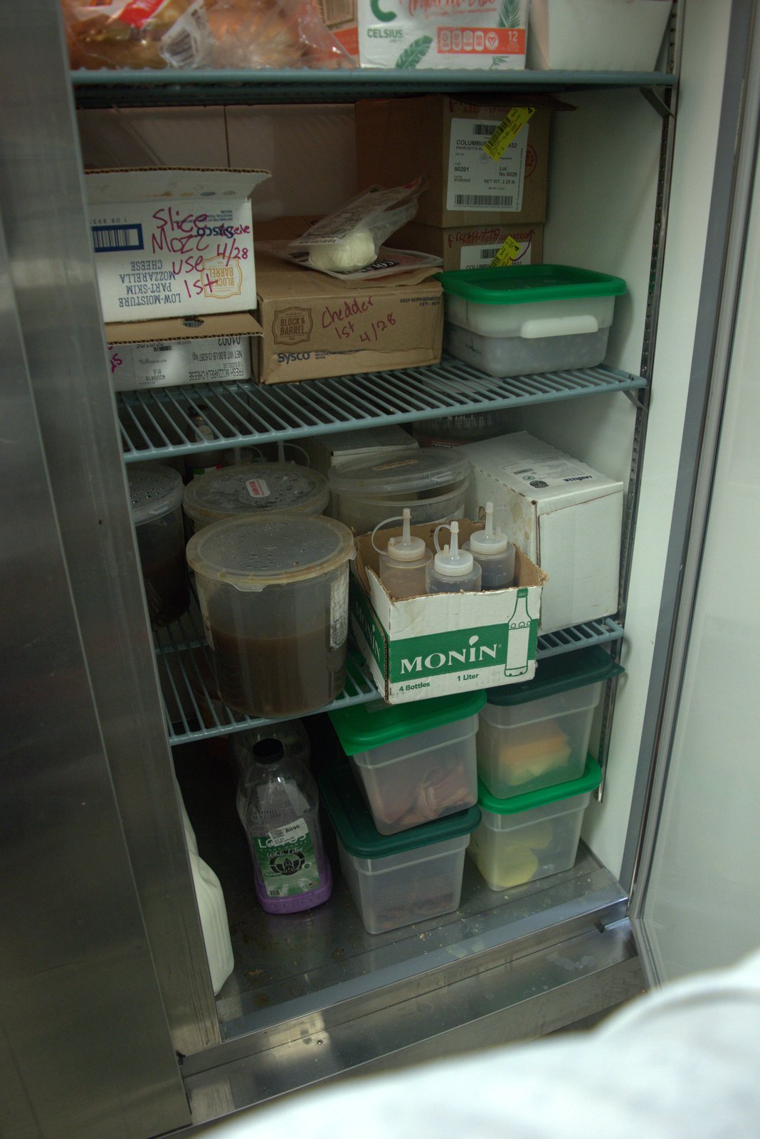 Same fridge, lower shelves — repurposed Monin shipping box used to hold uncovered squeeze bottles of sauces; sticky residue on container exterior