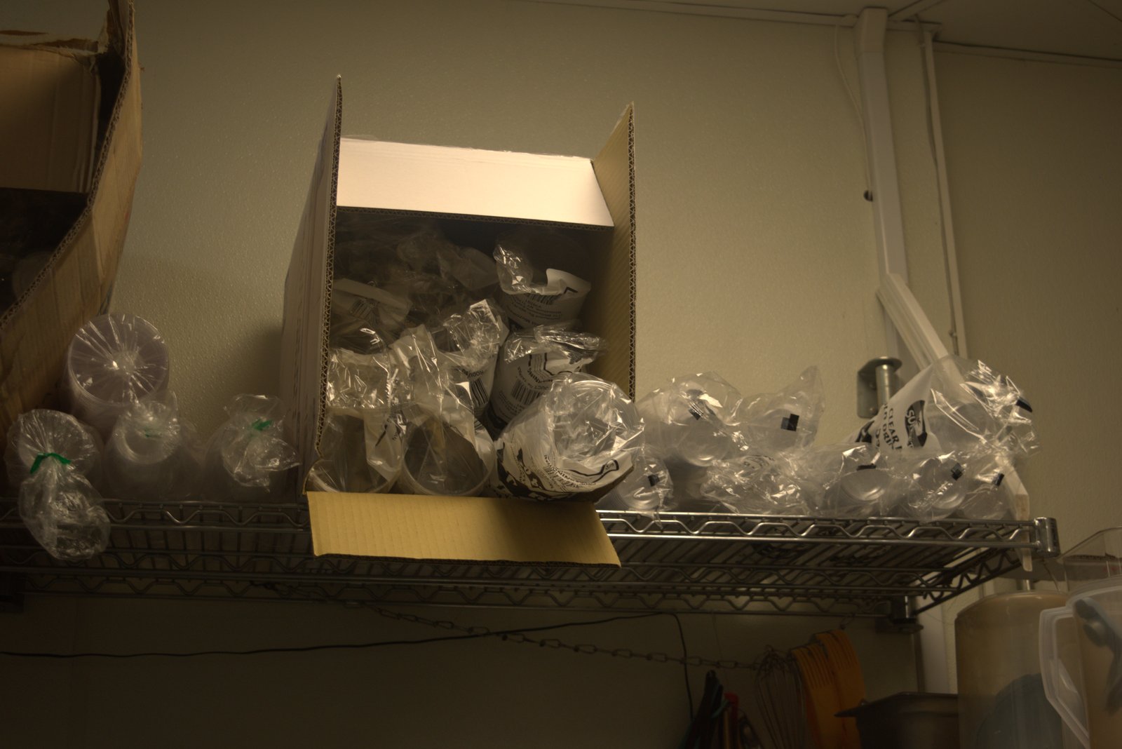 Overhead shelf above prep area — open box of unbranded plastic cups in loose plastic bags, no brand-approved packaging or seals
