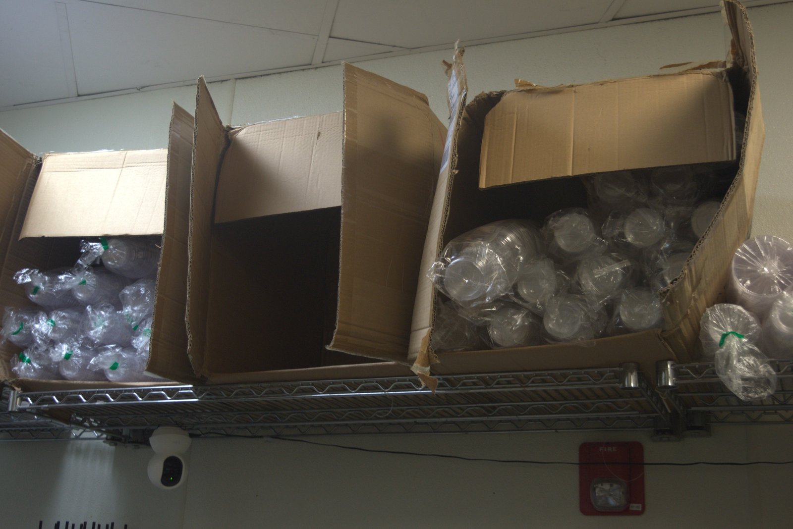 Top of overhead shelving — multiple torn cardboard boxes stuffed with bagged unbranded clear cups, dust accumulation around fire alarm