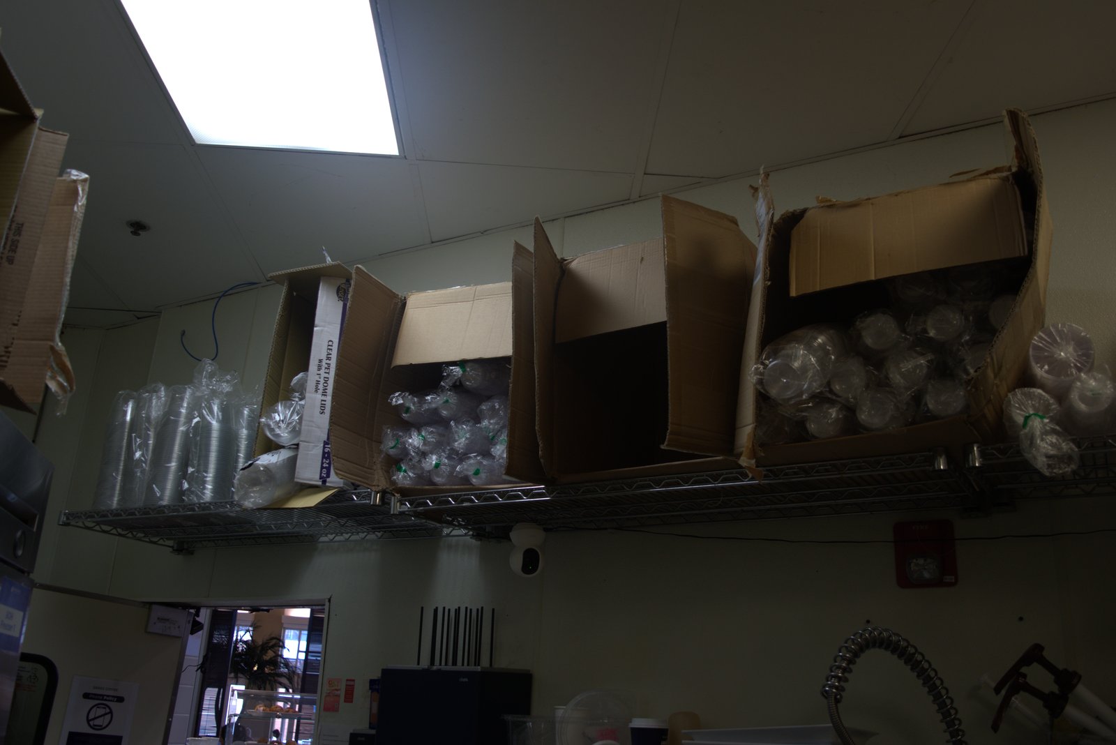 Wide view above the customer service area — torn boxes of unbranded cups and 'CLEAR PET DOME LIDS' stored on overhead racks, visible to customers through the open kitchen