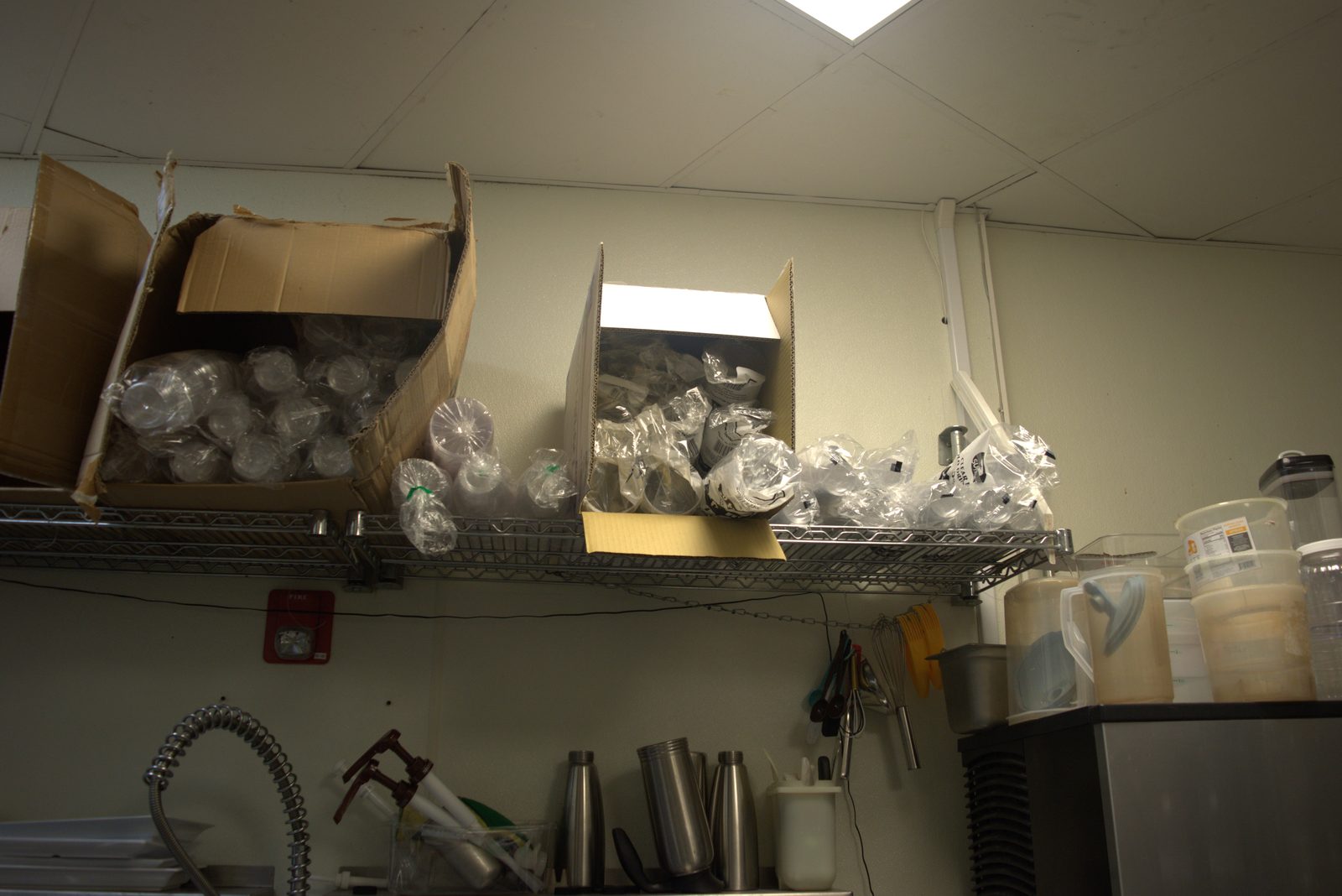 Overhead wire shelving — torn cardboard boxes stuffed with loose, bagged unbranded plastic cups directly above food-contact equipment