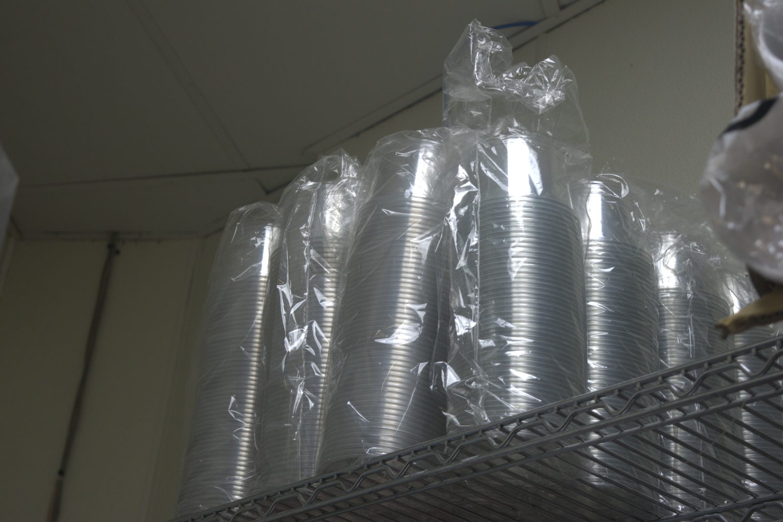 Close-up — bundles of unbranded clear plastic cups in loose, untied poly bags piled on overhead shelving, no Qargo-branded packaging, no protective covering