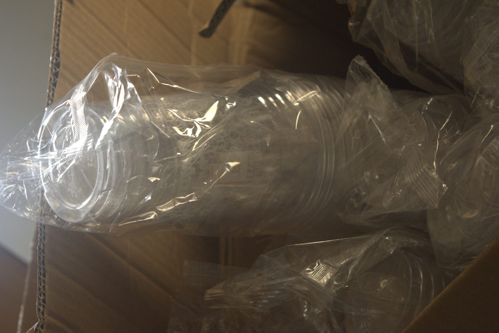 Bagged stacks of unbranded clear cups inside a torn cardboard box — exposed to dust and contamination from the surrounding stockroom
