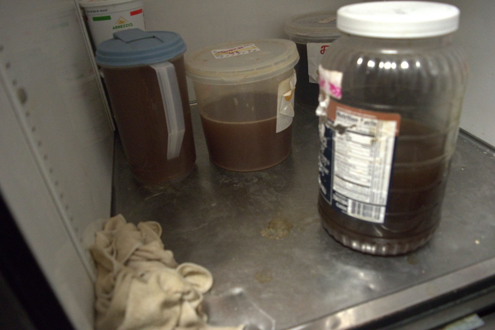 Bottom of the walk-in fridge — unlabeled containers of brown liquid and a soiled cloth lying directly on the fridge floor
