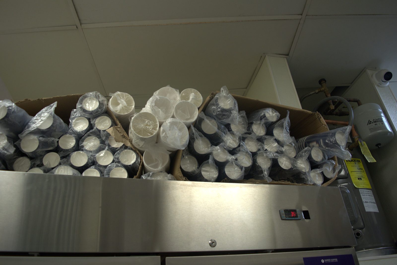 On top of the back-of-house refrigerator — torn cardboard boxes of bagged disposable cups exposed to ceiling dust