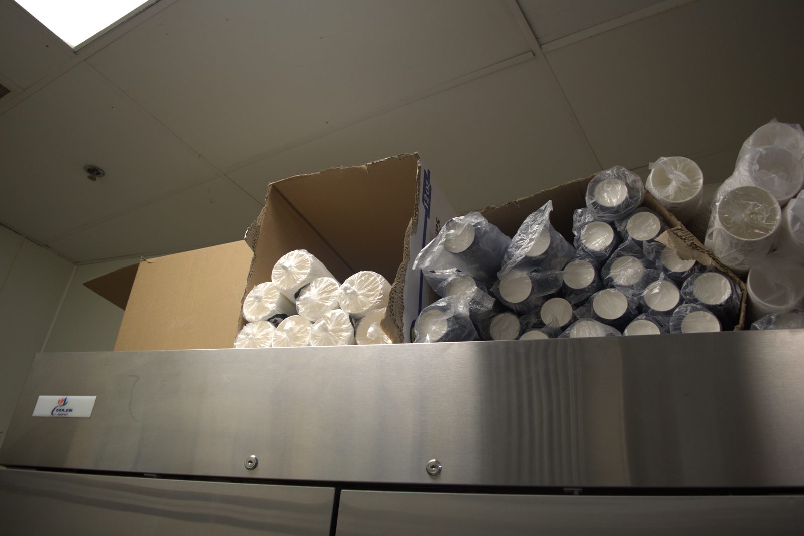 Closer view — torn boxes of bagged disposable cups stacked on top of the fridge ('Cooler Depot' unit), unsealed plastic bags exposed to airflow and ceiling dust