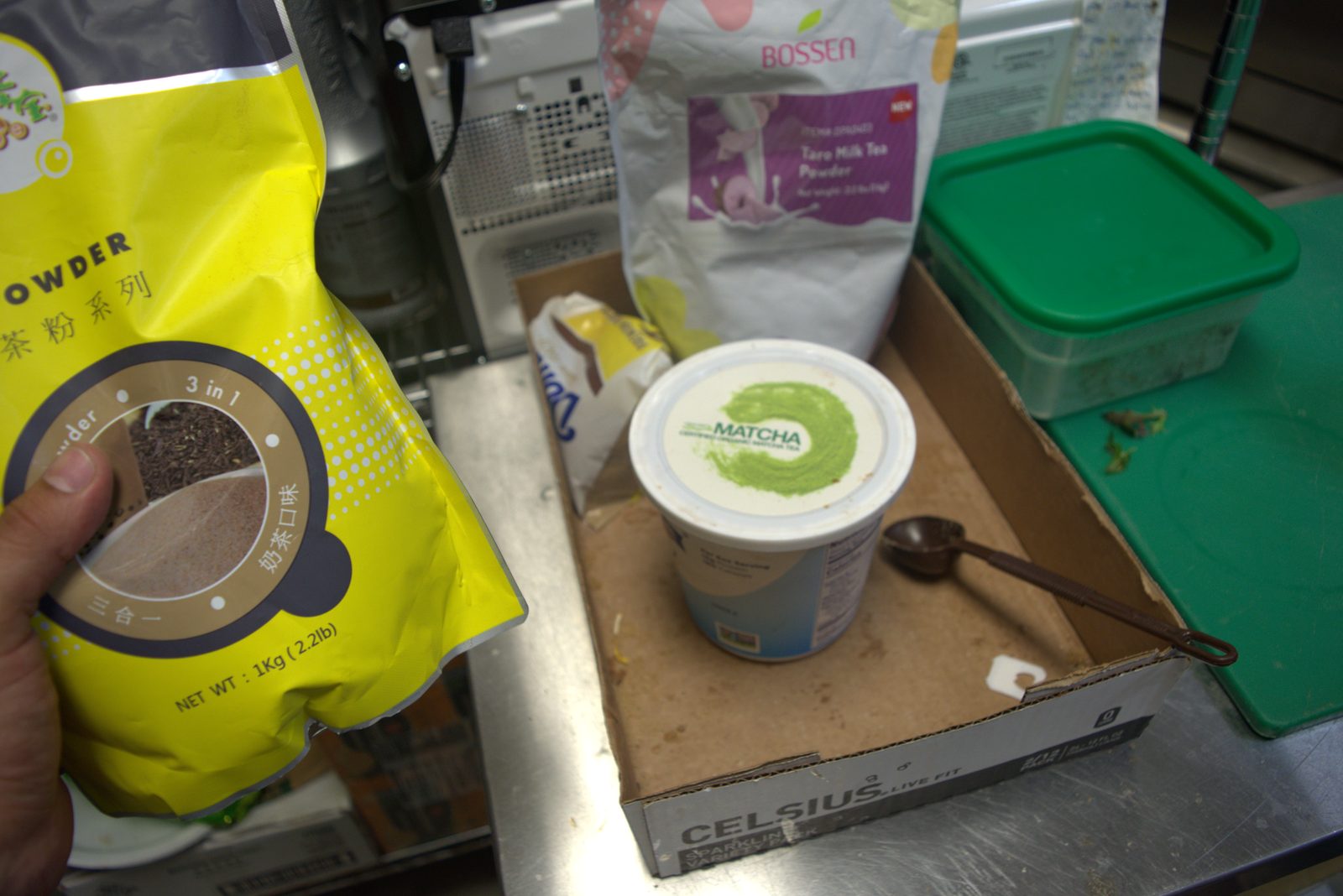 Chinese-language bulk milk-tea powder, Bossen Taro and Matcha sitting on a torn Celsius energy-drink shipping carton used as a tray