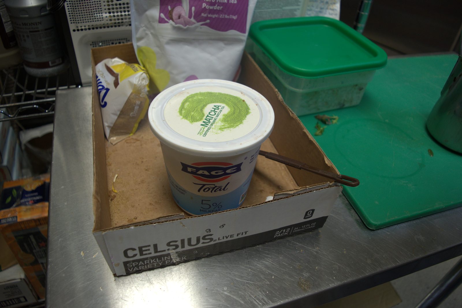 Retail Fage Greek yogurt tub being used as the ingredient container for matcha service, on a torn energy-drink box