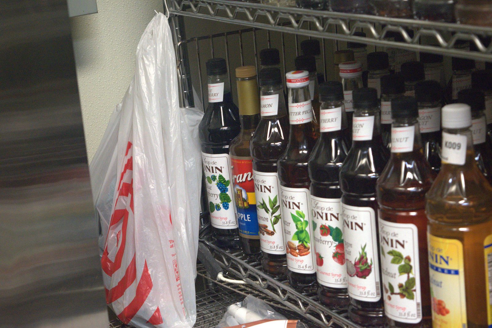 Same syrup shelving, closer view — dozens of Monin and Torani bottles confirmed by label; a white Target plastic bag is hung on the shelving, indicating retail grocery-store sourcing of supplies