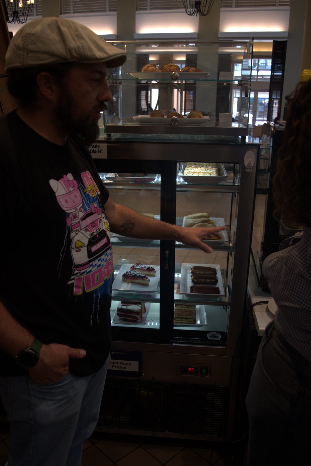 Wide view — operator gesturing at the 'Front Food Fridge' pastry case at 37°F; products on display include muffins, Napoleon, tiramisu, red velvet, and breakfast sandwiches with handwritten marker labels directly on the glass