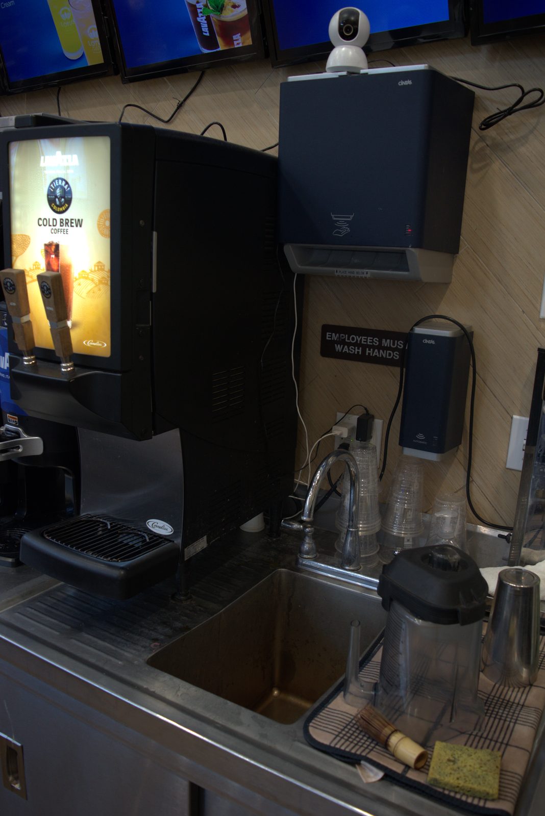 Lavazza-branded Cold Brew Cornelius dispenser at the service counter; consumer Wi-Fi webcam zip-tied above the hand-wash dispenser