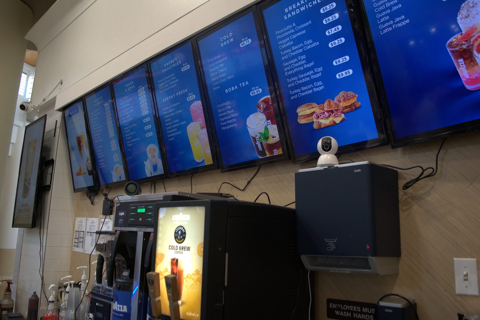 BOH freezer area — handwritten whiteboard staff schedule mounted next to the 'BOH Freezer 1' (required corporate scheduling system not in use); a consumer Wi-Fi pan/tilt webcam is zip-tied to the menu-board frame above the schedule, again replacing the required Solink camera