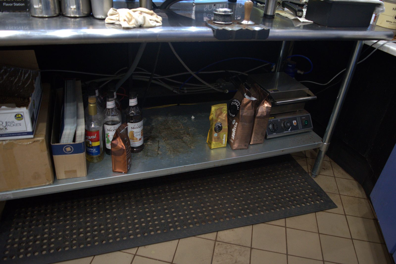 Under-counter shelf at the espresso station — Lavazza coffee bags and unauthorized Monin/Torani syrups as the active backup stock