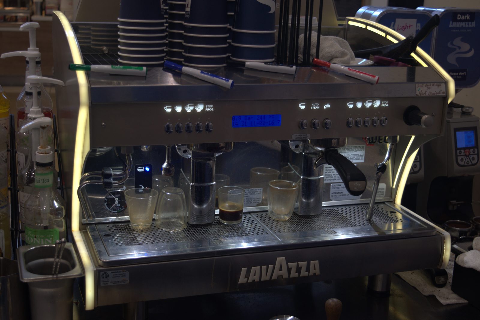 Same Lavazza espresso machine, wider view — flanked by Monin syrup bottles (Thai Tea, Coconut, etc.) on a side rack; cup warmer cluttered with marker pens and rags directly above stacked clean service cups