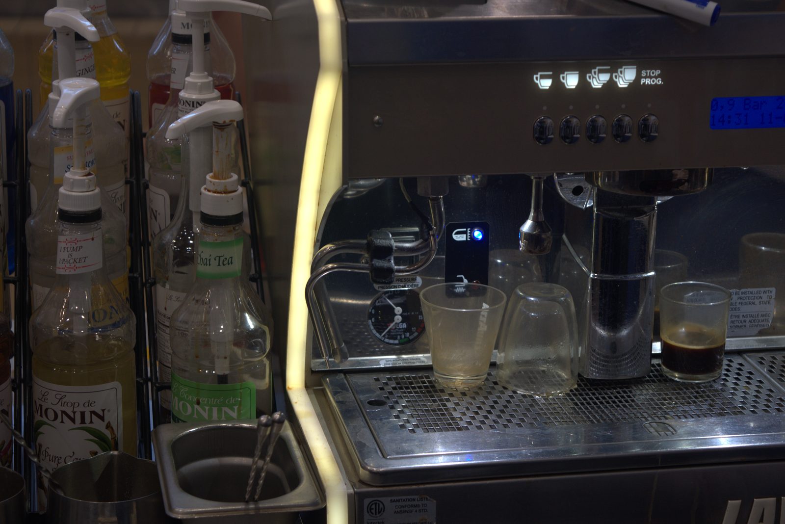 Detail at the espresso station — open Monin syrup pump bottles (Ginger, Sugar Free Sweetener, Thai Tea, Pure Cane) with sticky residue on caps, sitting next to the active Lavazza espresso group with used shot glasses on the drip tray