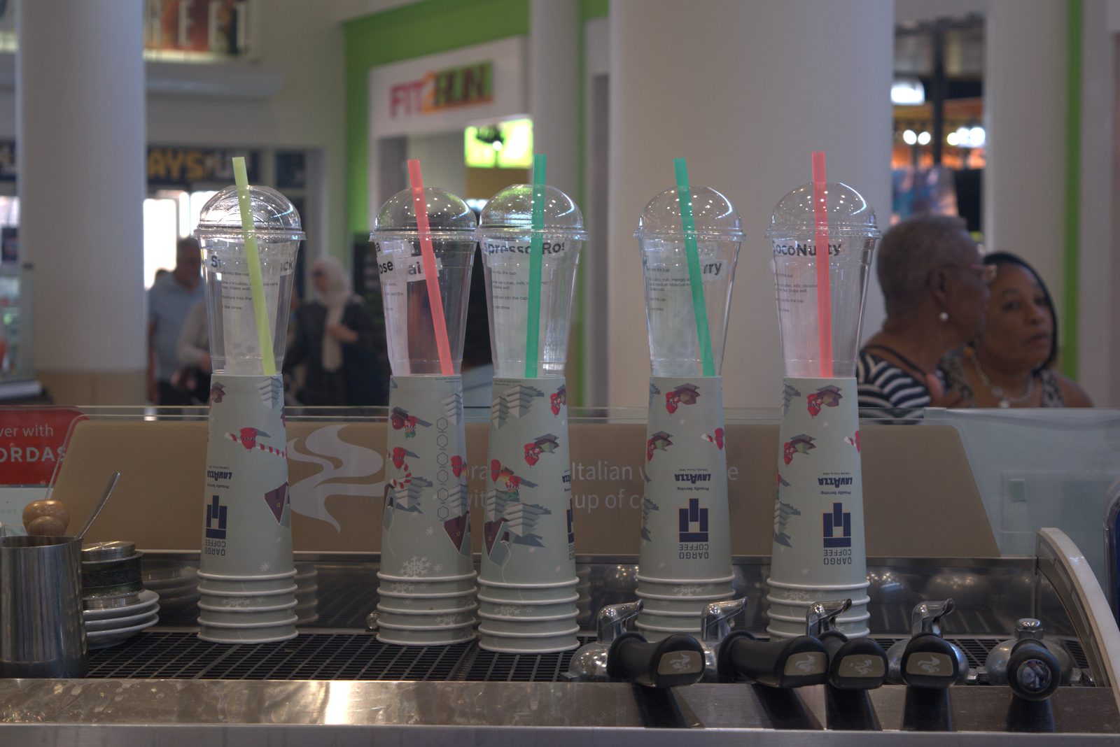 Same FOH counter, alternate angle — five drinks (Strawberry, Espresso Rose, CocoNutty, etc.) all served in 'Qargo Coffee — Proudly Serving LAVAZZA' cups in front of customers in the mall food court