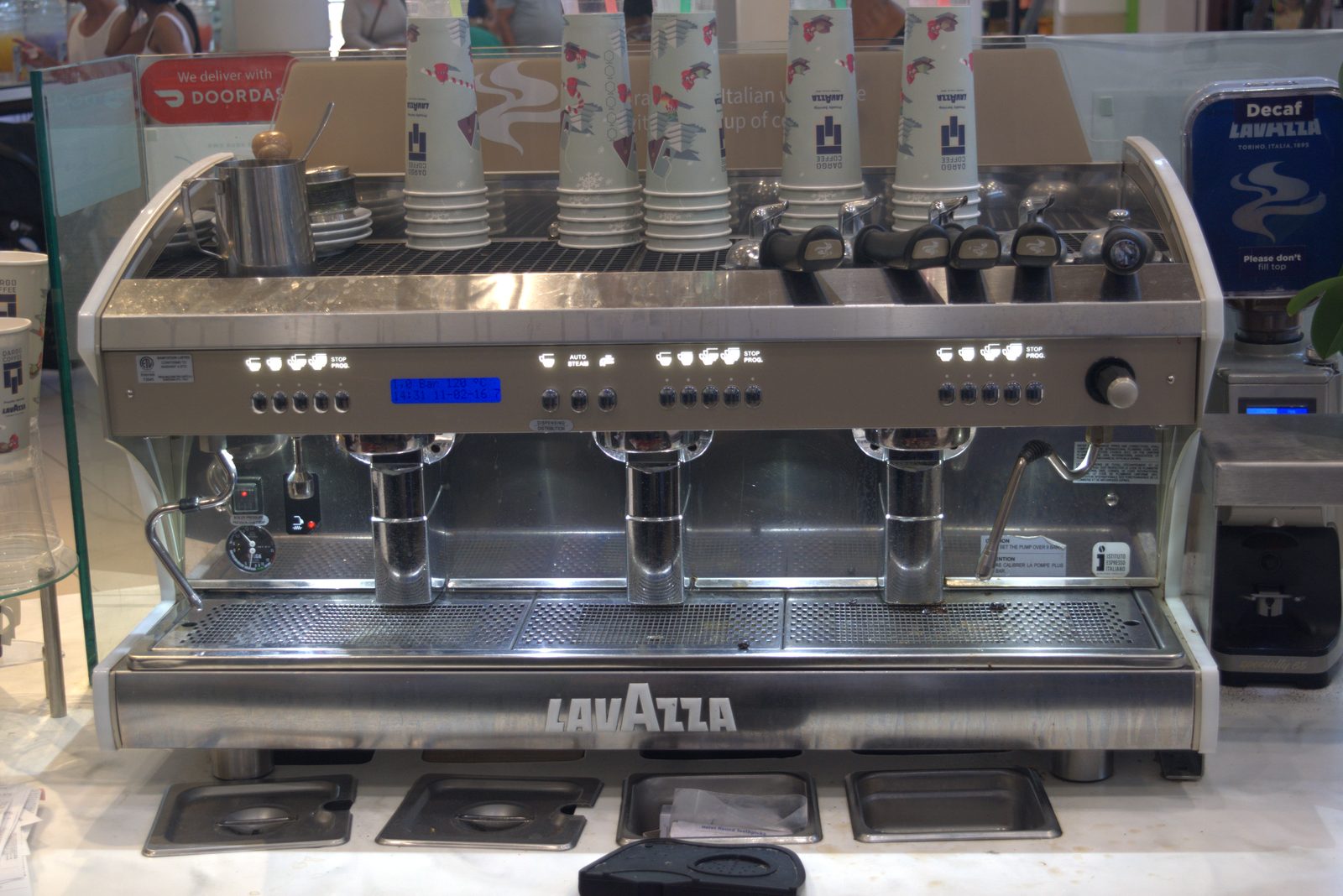 Wider view of the same Lavazza three-group machine plus a Lavazza Decaf grinder on the right — entire espresso program is Lavazza-branded equipment and supplies, with a DoorDash decal on the side glass